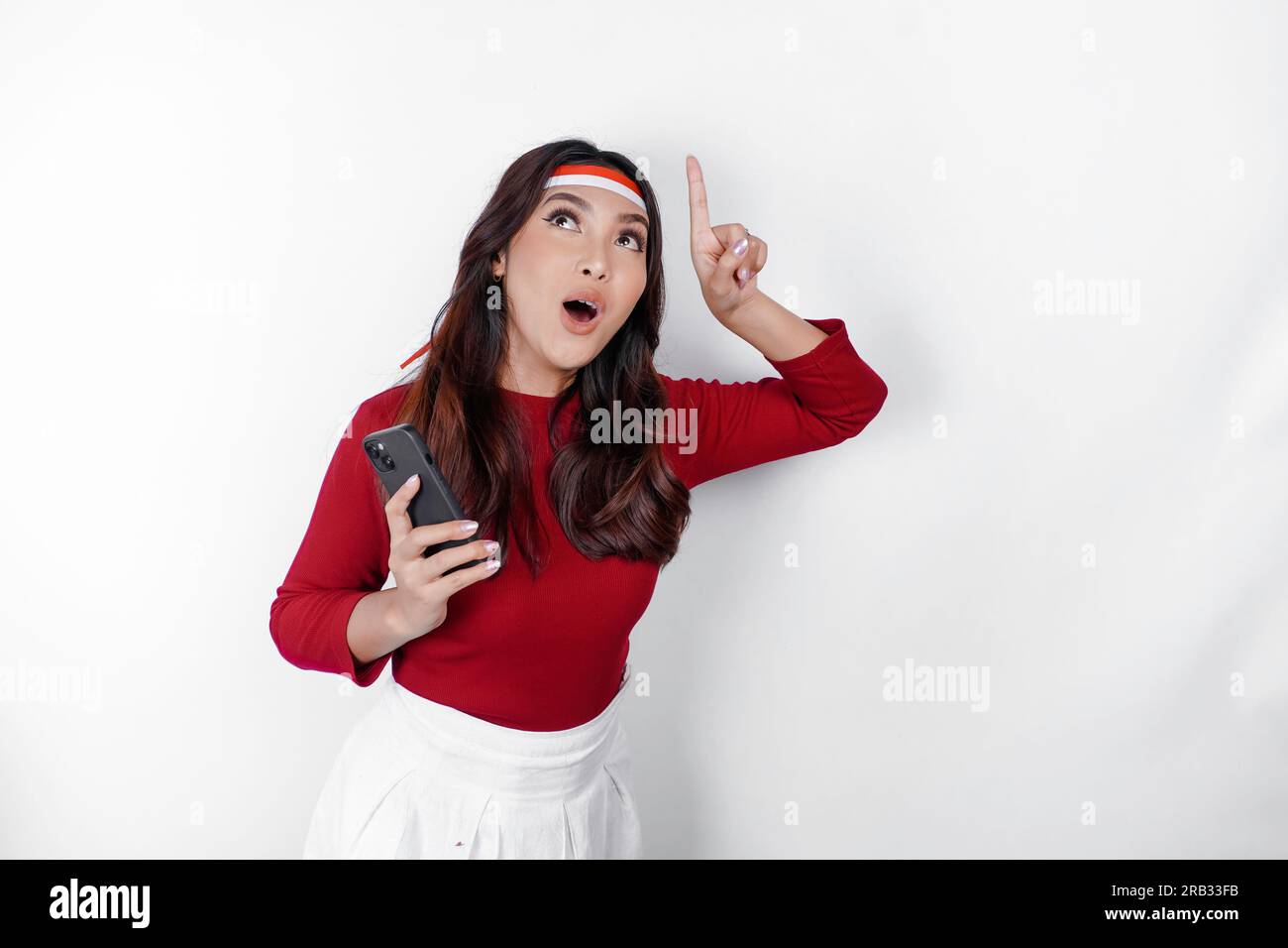 A portrait of a shocked Asian woman wearing headband, holding her phone, and pointing copy space on top of her, isolated by white background. Indonesi Stock Photo