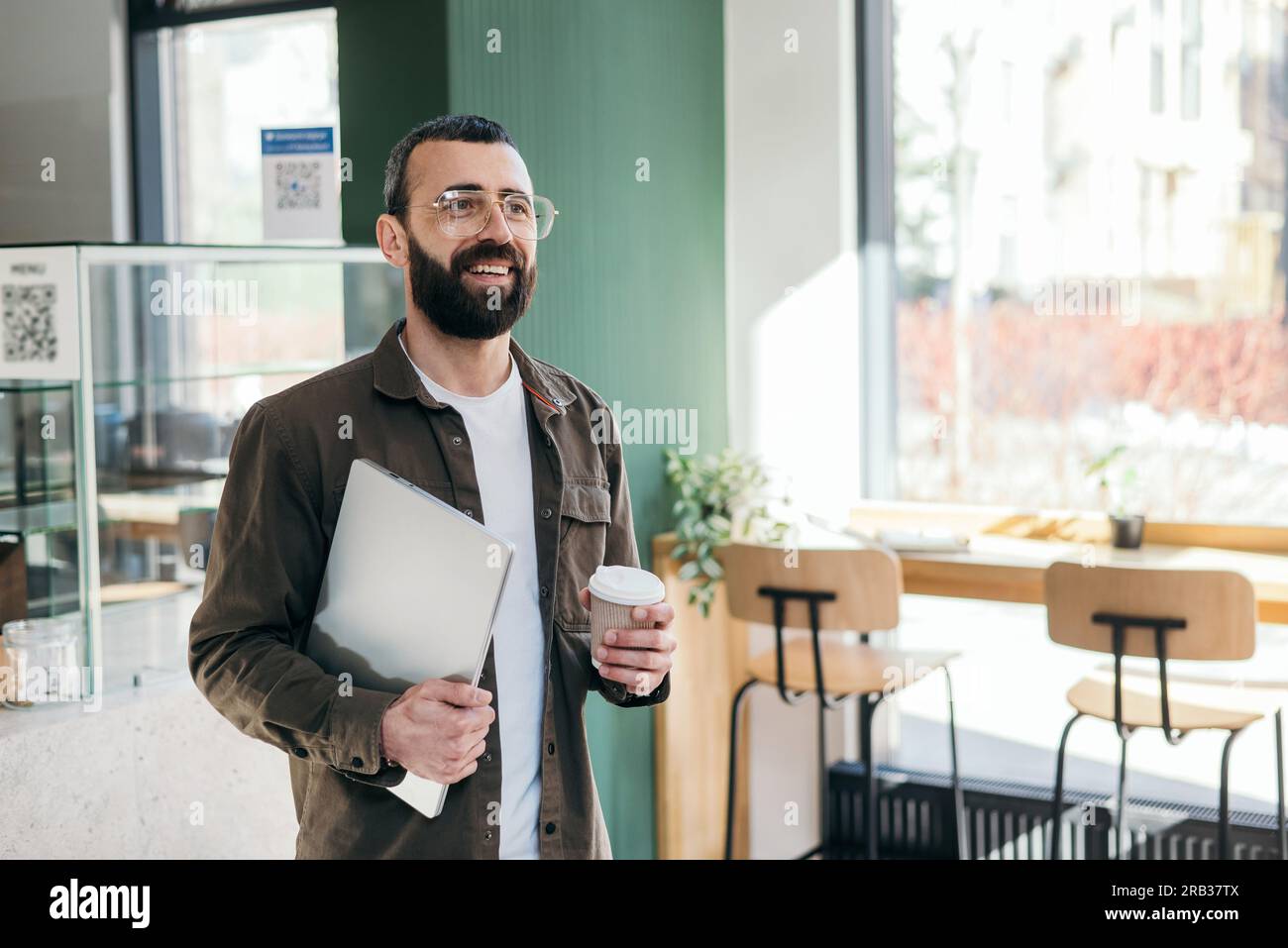 Smiling bearded creative man in glasses standing holding laptop and cup of coffcee in cafe looking out window. Freelancer and modern interior Stock Photo