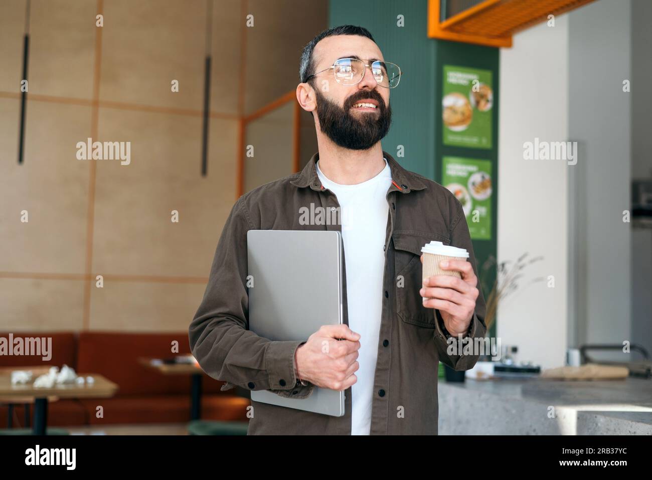 Smiling bearded creative man in glasses standing holding laptop and cup of coffcee in cafe looking out window. Freelancer and modern interior Stock Photo