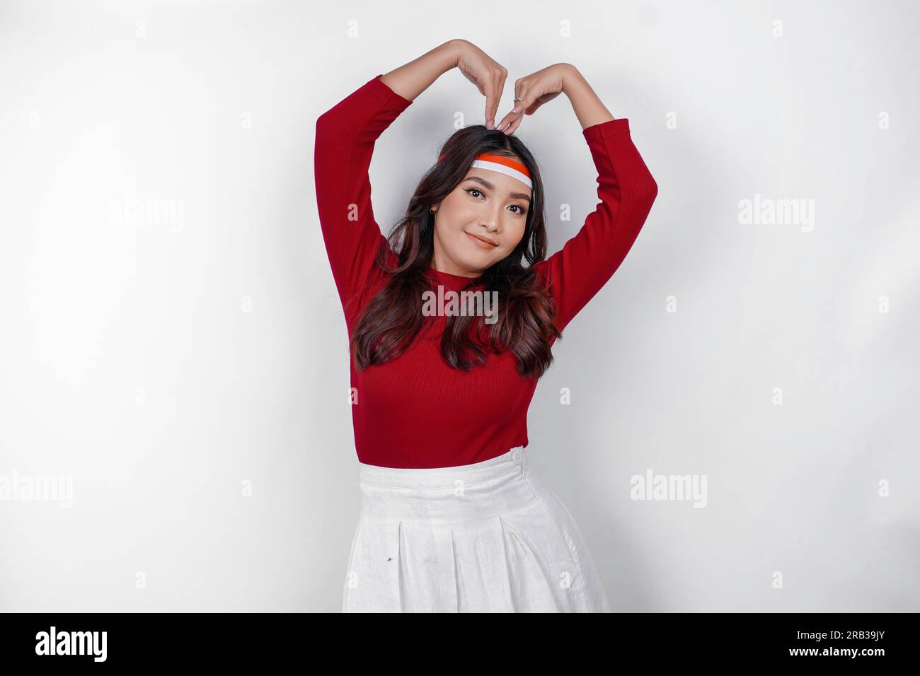 A young Asian woman wearing a red top feels nationalism, shapes heart gesture expresses her love for Indonesia. Indonesia's independence day concept. Stock Photo