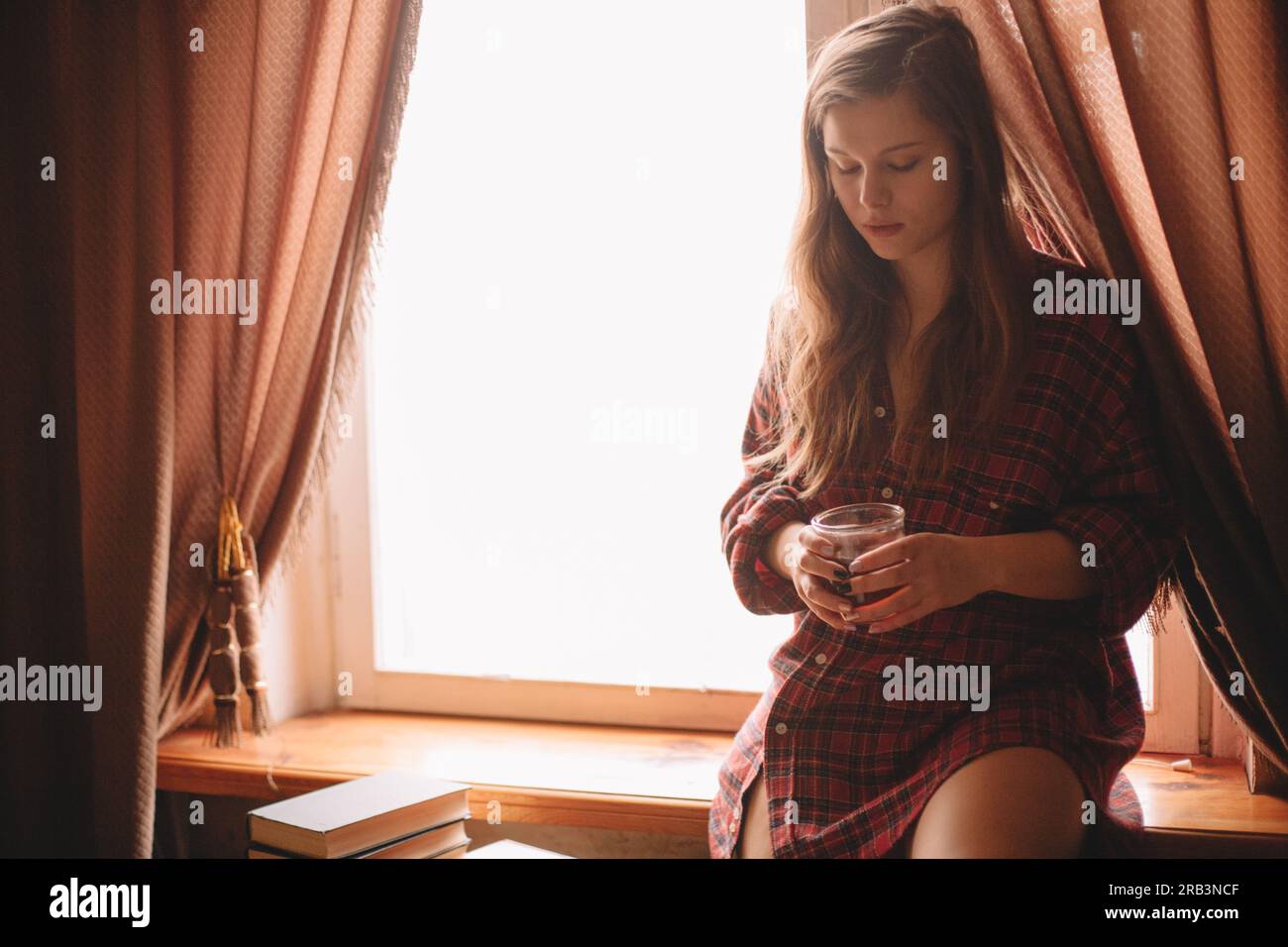 Young woman holding cup of tea while sitting on window sill Stock Photo