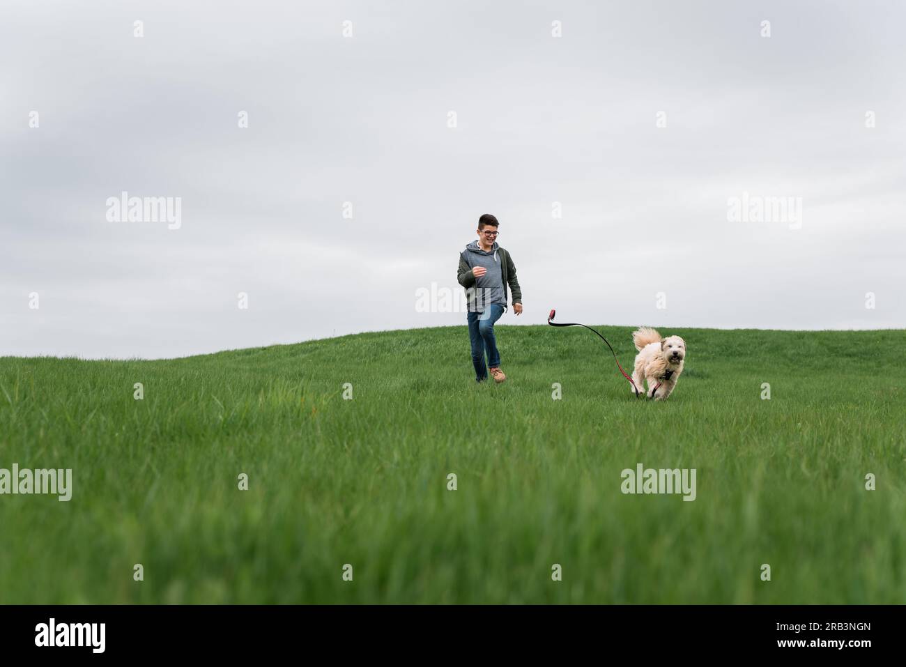 Teenage boy running across a grassy field with his dog on cloudy day. Stock Photo