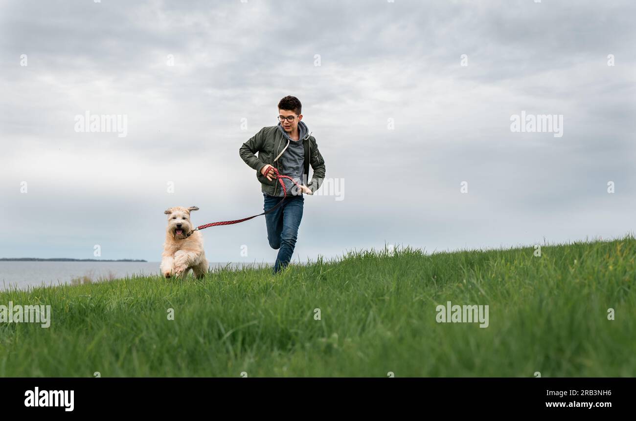 Teenage boy running up a grassy hill with his dog on cloudy day Stock