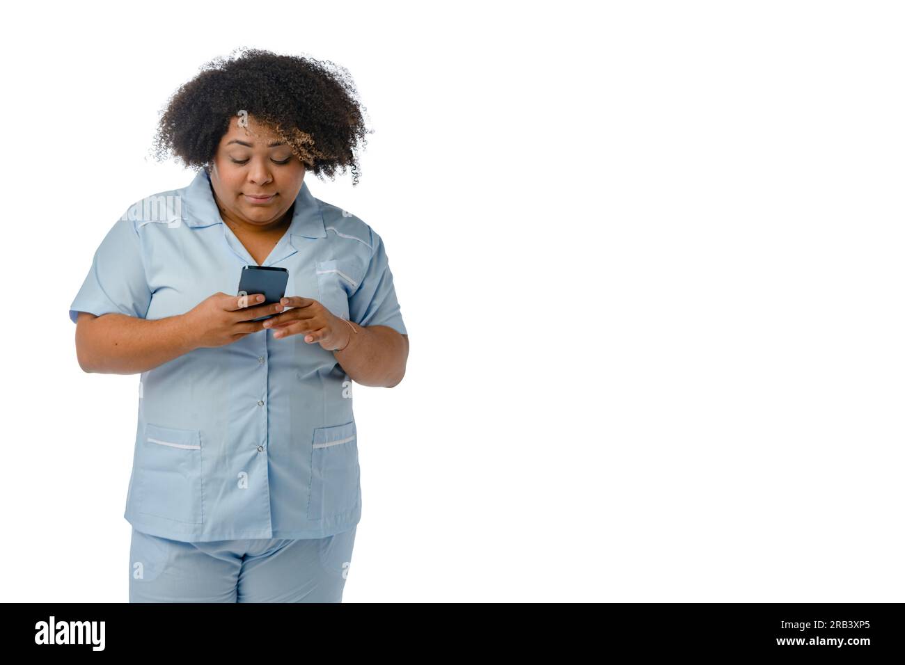 young Afro-Latin female doctor in blue uniform, is standing watching medical appointments on the phone, reading messages and notifications, white back Stock Photo