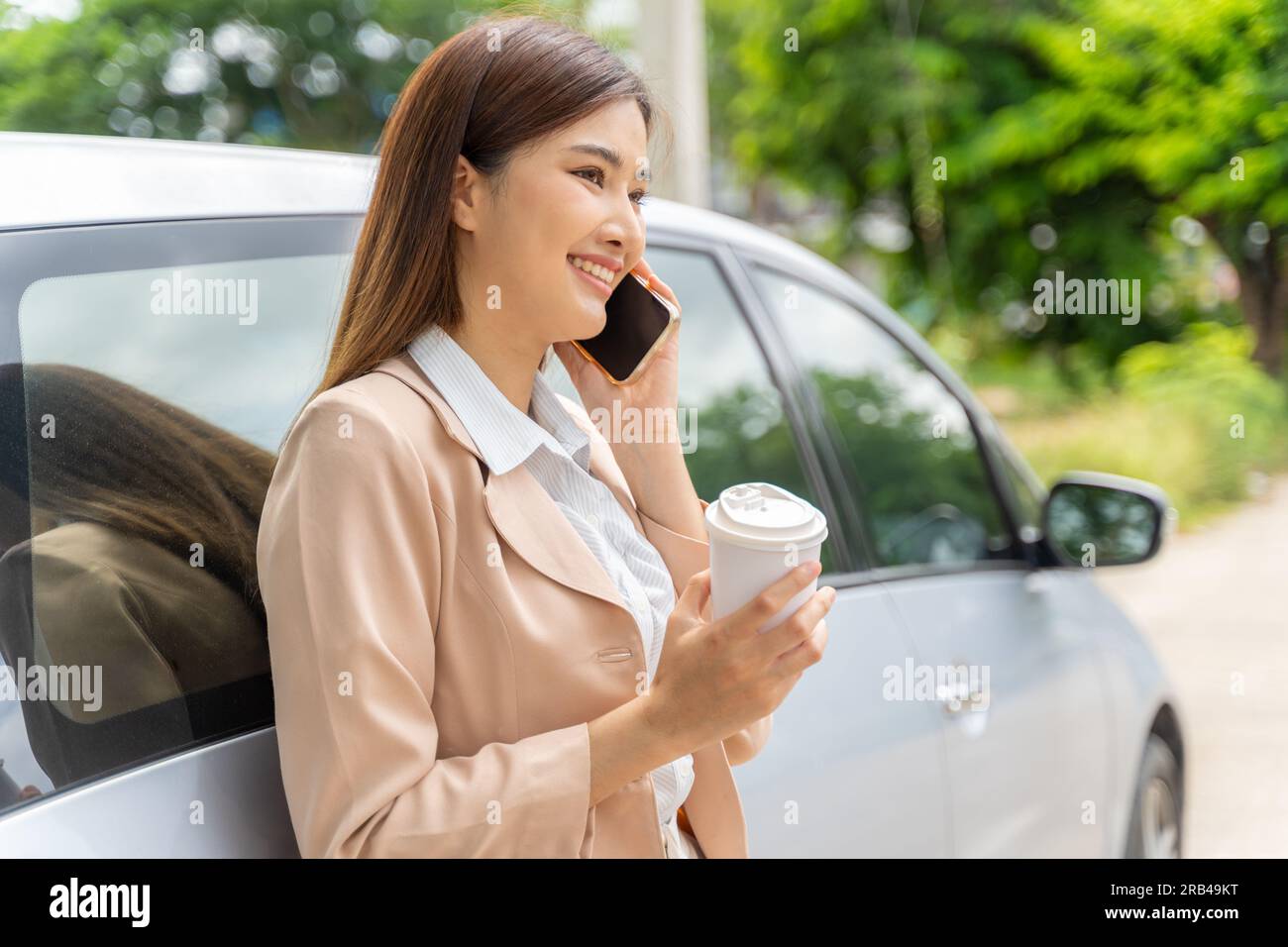 Happy smiling young working woman holding a take away cup of coffee while talking on her phone and leaning against her car Stock Photo