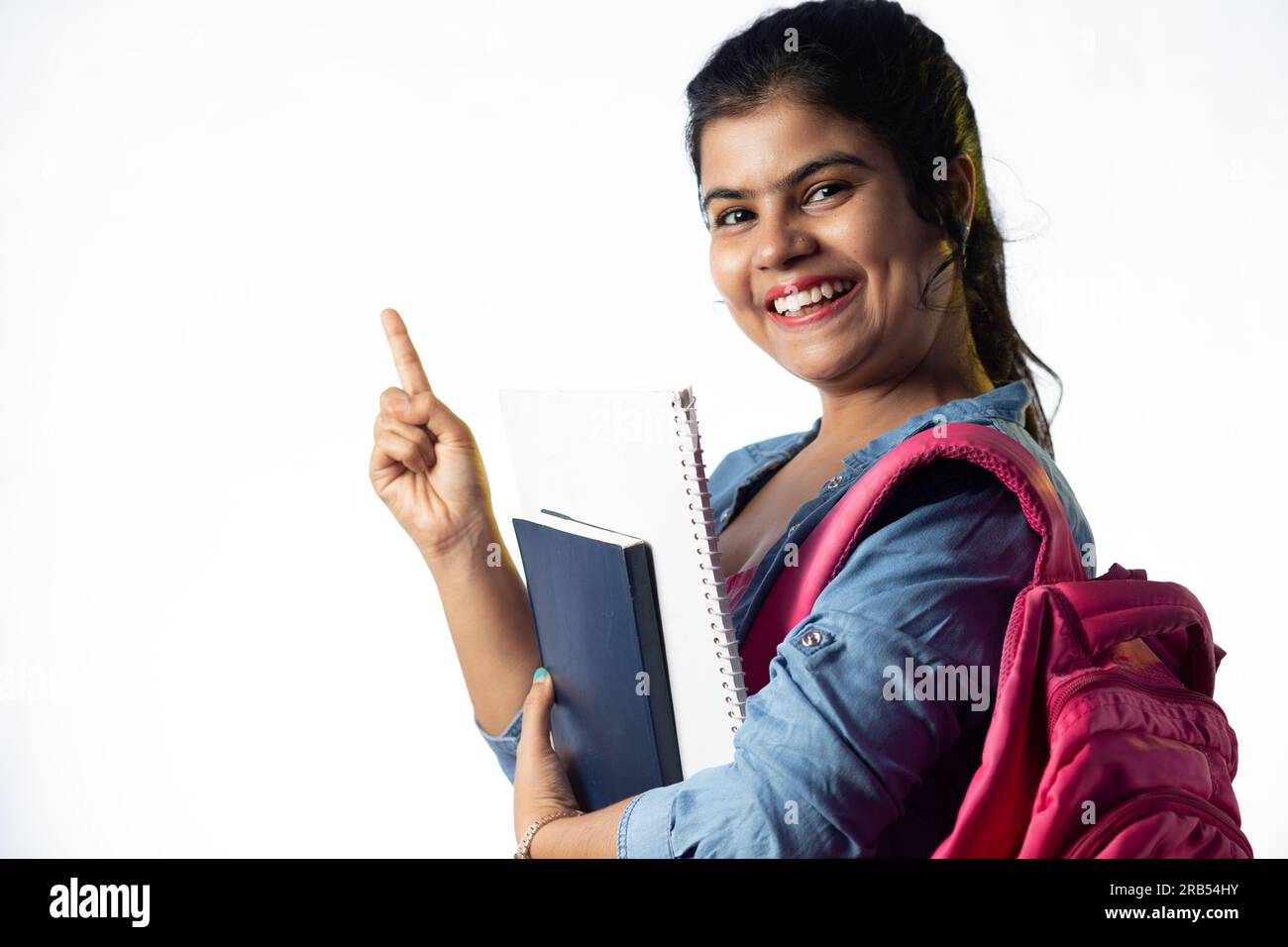 A pretty young Indian girl with books in hand and backpack pointing ...