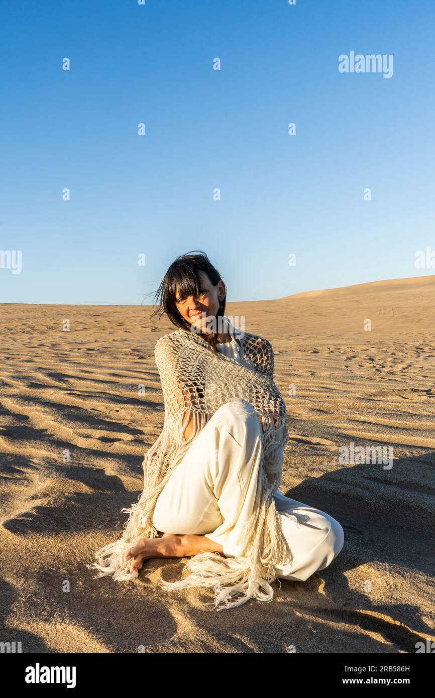 A woman dressed in white sitting on the sand at sunset Stock Photo