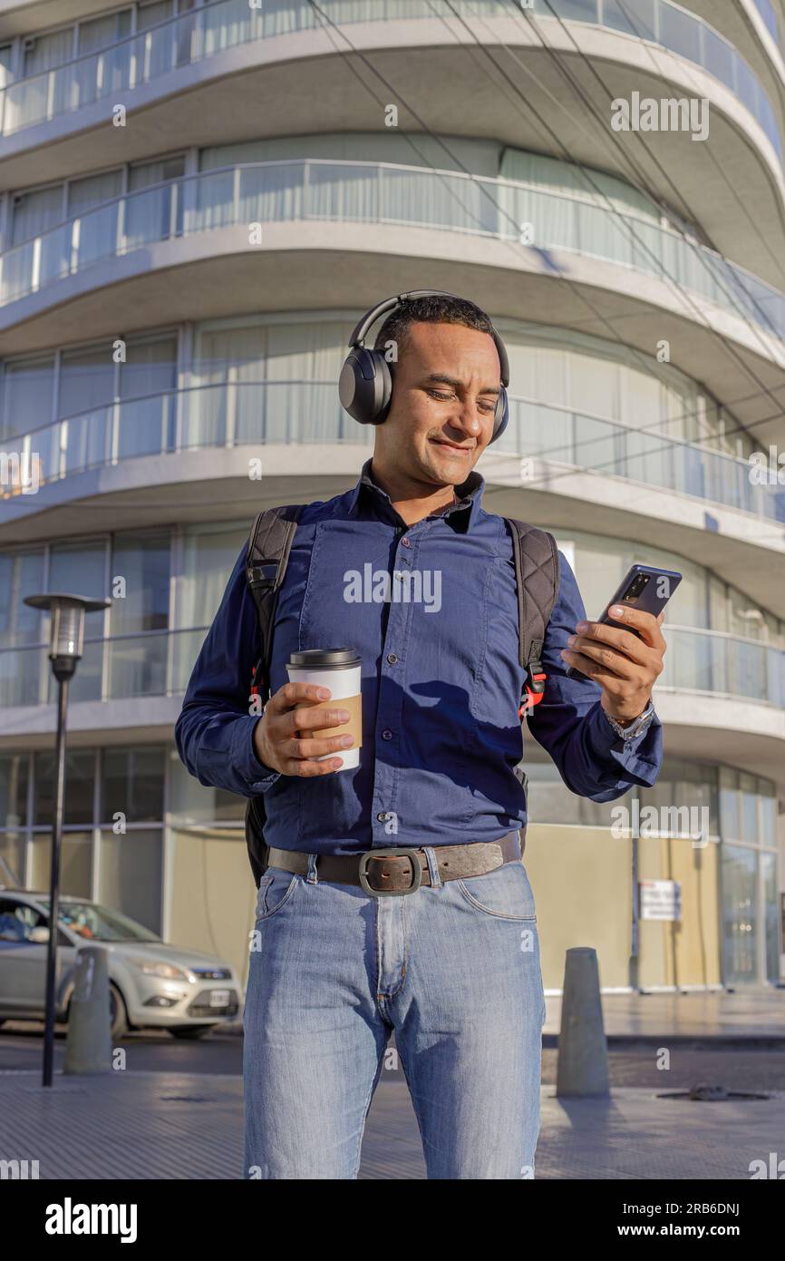 Smiling young latin man with headphones looking at his mobile phone and holding a paper cup with coffee in his hand. Stock Photo