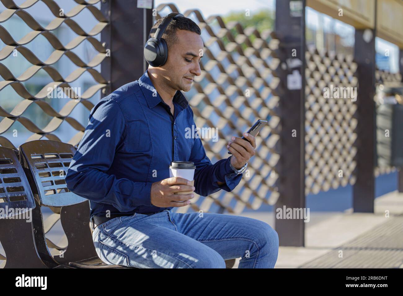 Young latin man with headphones sitting waiting for the bus with a paper cup of coffee in his hand. Stock Photo
