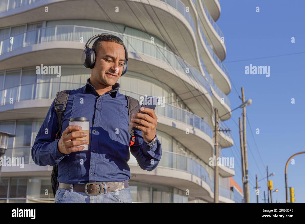 Young latin man with headphones looking at his mobile phone and holding a paper cup with coffee in his hand. Stock Photo