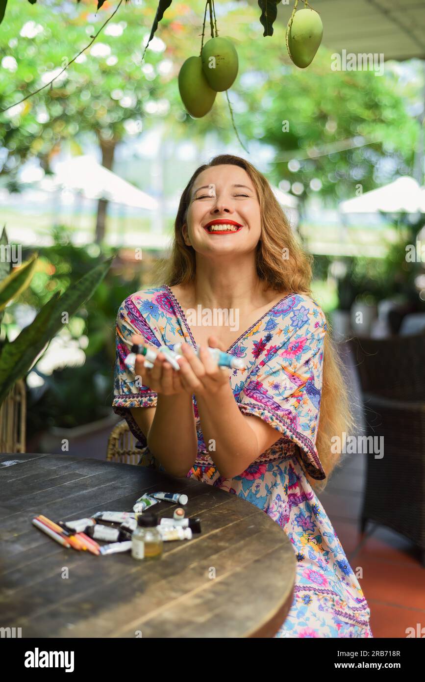 Portrait of beautiful blonde woman artist and illustrator  sitting by the table and happily holding her acrylic paints Stock Photo