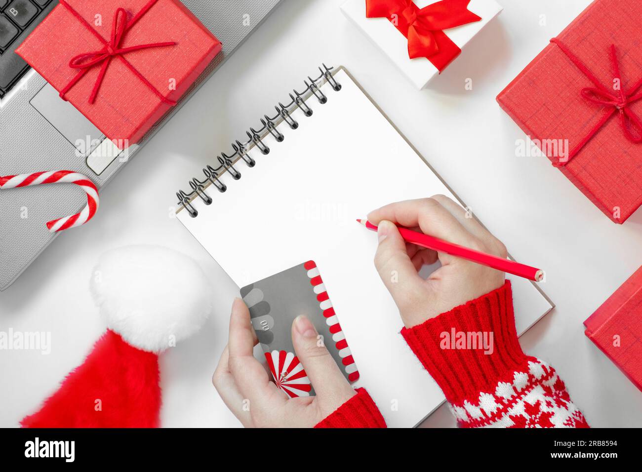 Christmas online shopping flat lay. Girl writing shopping list. Female Hands with Credit card and Pencil. Laptop and christmas presents on white table Stock Photo