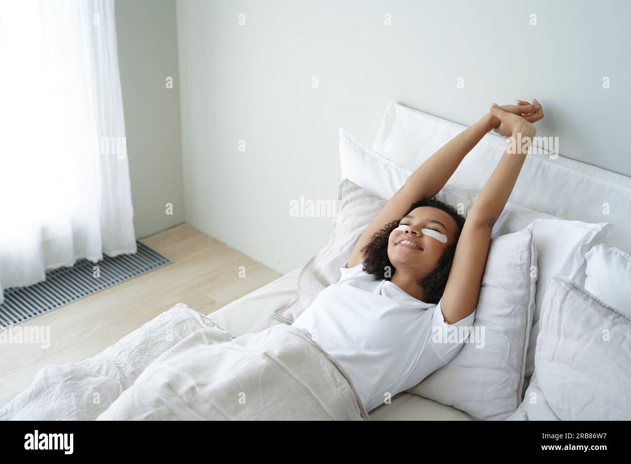 Biracial teen stretches in bed, wearing under-eye patches. Happy girl enjoys skincare, morning beauty routine. Stock Photo