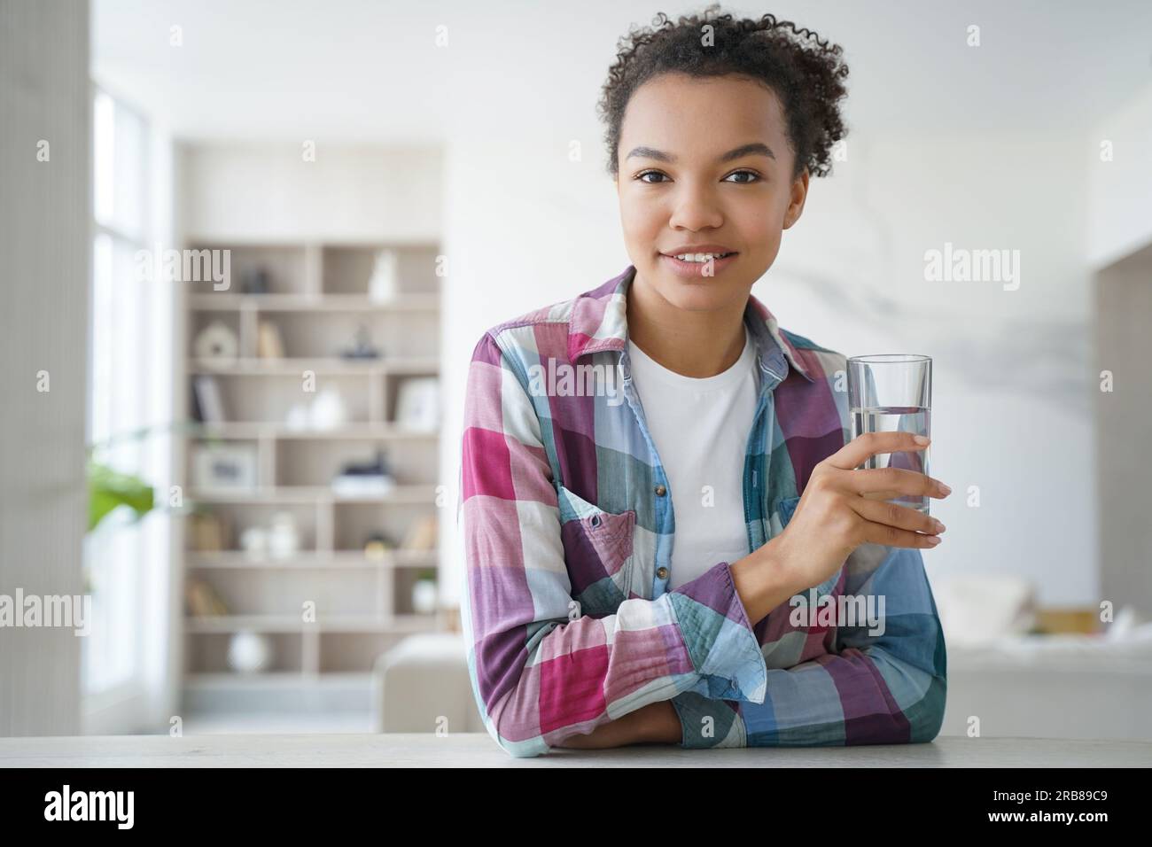 Happy biracial girl at home holds glass of water, promoting healthy lifestyle, smiles for camera. Stock Photo