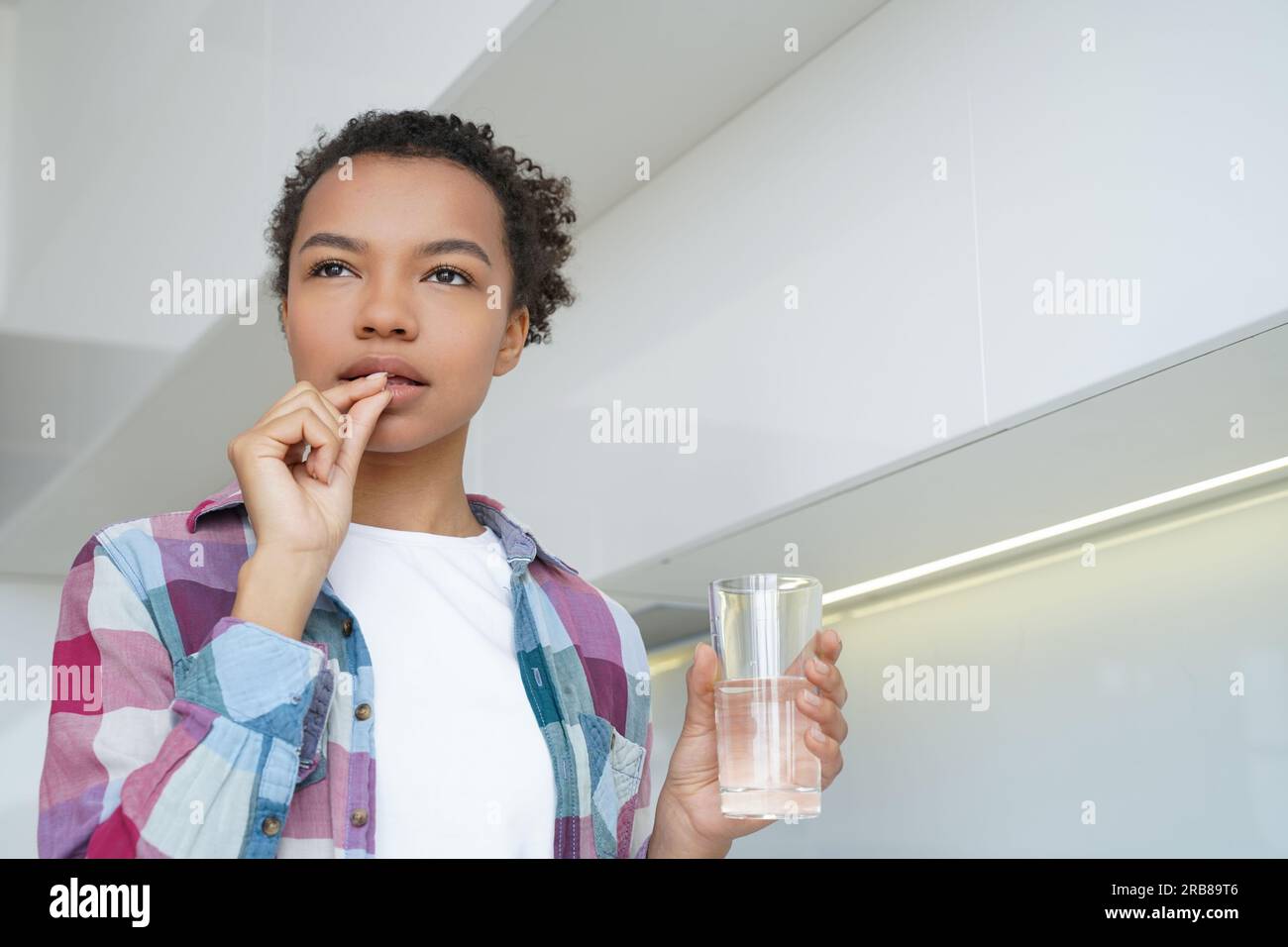 Pensive biracial girl takes pill, holding water, promoting healthy lifestyle and wellness with medicine, supplements, vitamins. Stock Photo