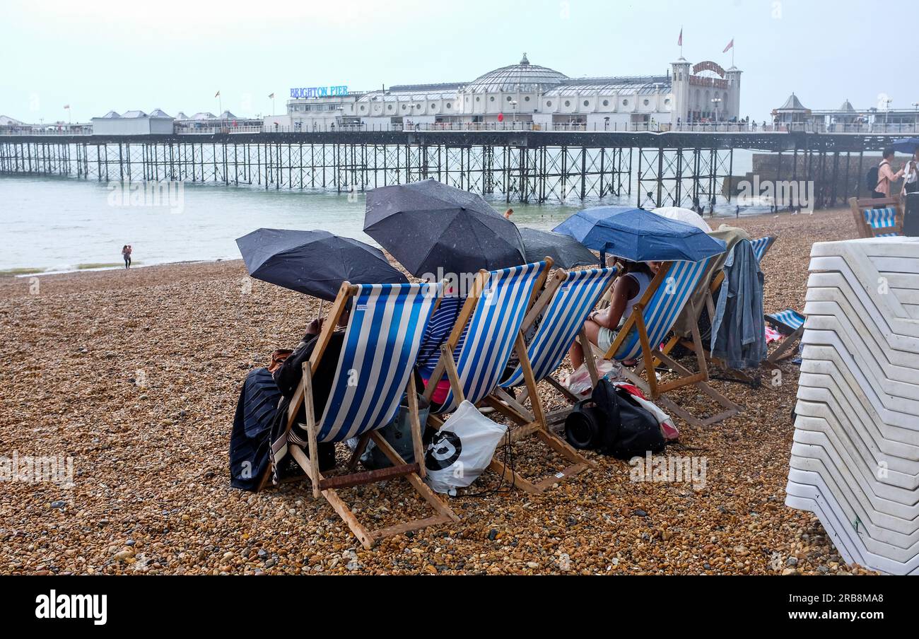 Brighton UK 8th July 2023 - Visitors try to stay dry on Brighton beach under umbrellas during torrential rain showers  as thunder storms sweep across parts of the UK today  : Credit Simon Dack / Alamy Live News Stock Photo