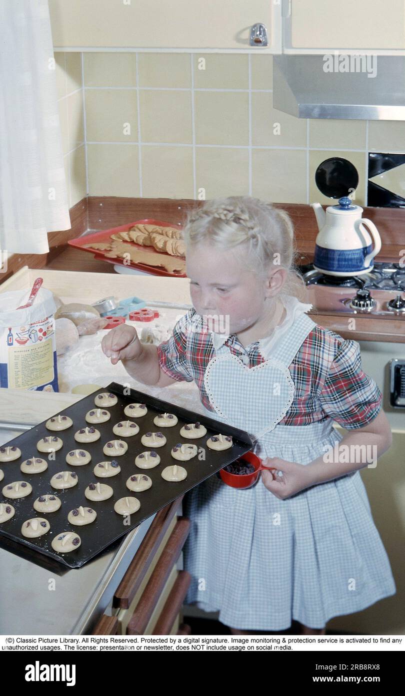 In the early 1960s. A girl nicely dressed in an apron puts raisins on top of the almost ready to bake cookies. It looks like it's close to christmas time as there is already a tray of ready made gingerbread on the counter. The kitchen interior is typical of the decade. Sweden 1960 Stock Photo
