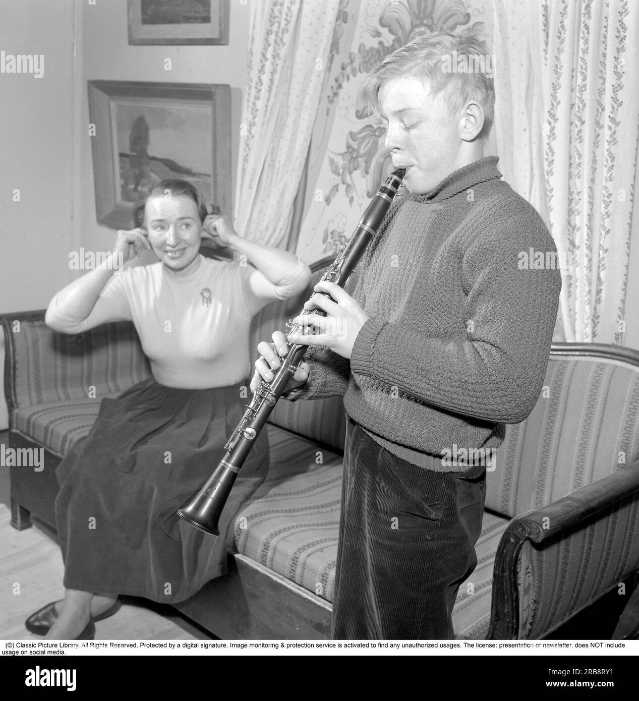 At Rune Lindström's home (1916-1973)  Swedish screenwriter and actor.  Here his wife with their son in a picture taken in their home in Leksand in 1958. Sweden. He is seen happily playing his clarinet thinking it sounds good but judging by his mother's reaction and her fingers in her ears as if to protect herself from the noise, the playing does not sound good. Conard ref 3672 Stock Photo
