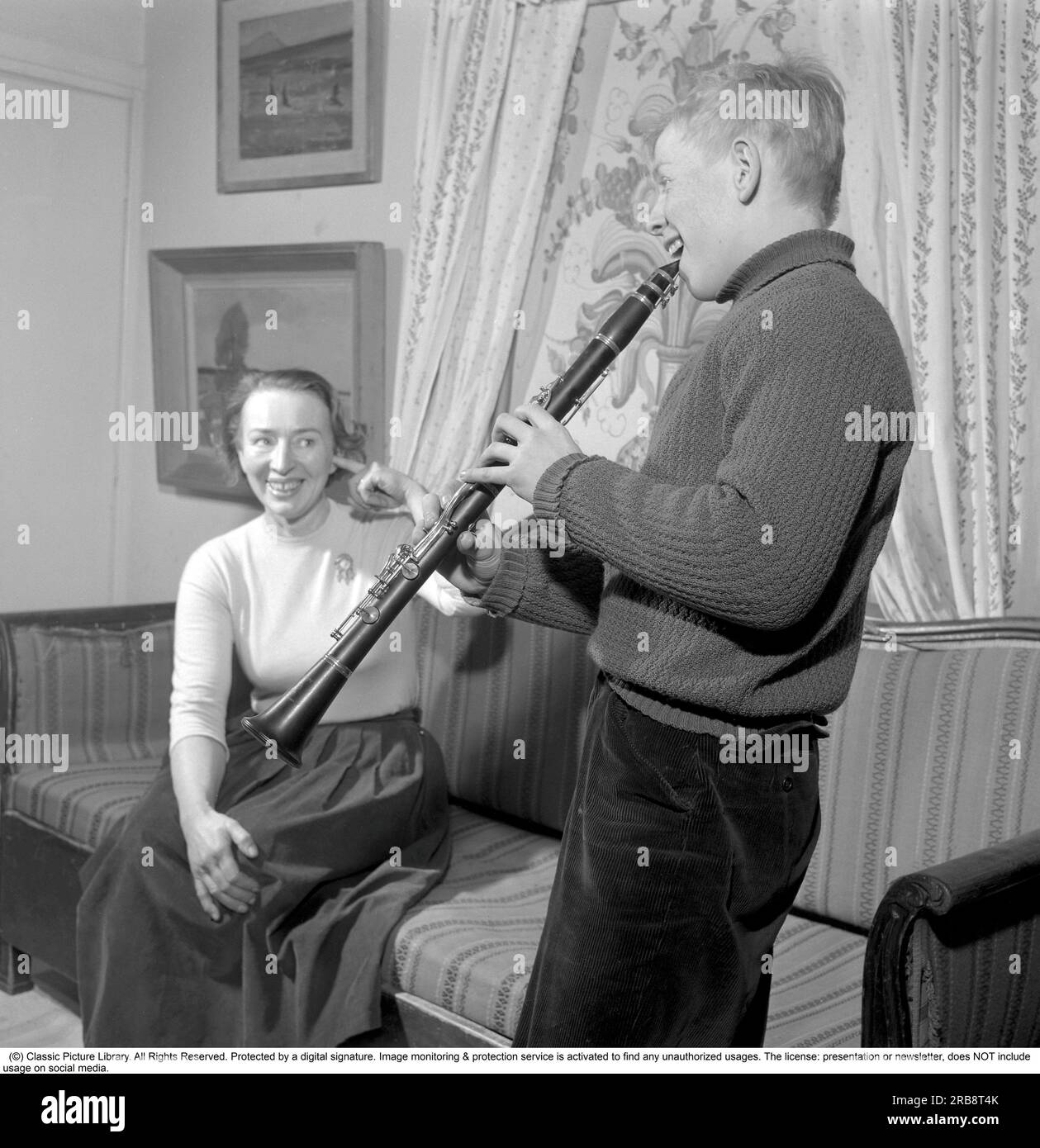 At Rune Lindström's home (1916-1973)  Swedish screenwriter and actor.  Here his wife with their son in a picture taken in their home in Leksand in 1958. Sweden. He is seen happily playing his clarinet thinking it sounds good but judging by his mother's reaction and her finger in one ear, as if to protect herself from the noise, the playing does not sound good. Conard ref 3672 Stock Photo