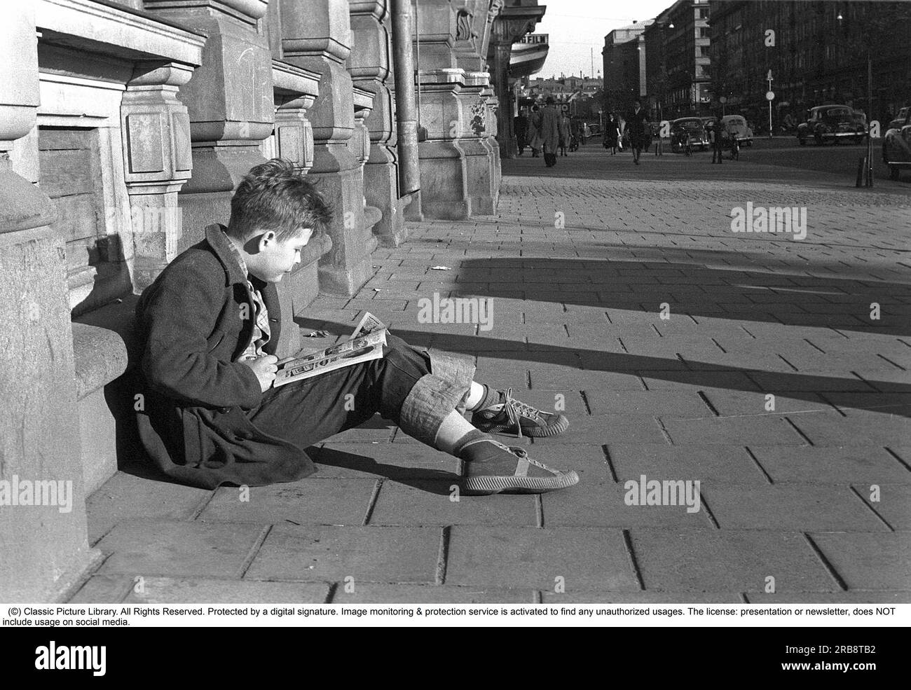 Stockholm in the 1950s. A boy sits outside Bonnier's publisher's building at Sveavägen 56 and reads a comic book. The year is 1953. Cinema Riviera can be seen a little further down the street Sveavägen 52. Farthest to the right is the Konserthuset at the intersection of Kungsgatan. With Kalle Anka & C:o and Seriemagasinet starting to come out in 1948, the comic book phenomenon took off in earnest in Sweden. After a boom in the 1950s, the development slowed somewhat, partly because the American series debate also reached Sweden. During the 1950s and 60s, comic books began to be considered 'inap Stock Photo