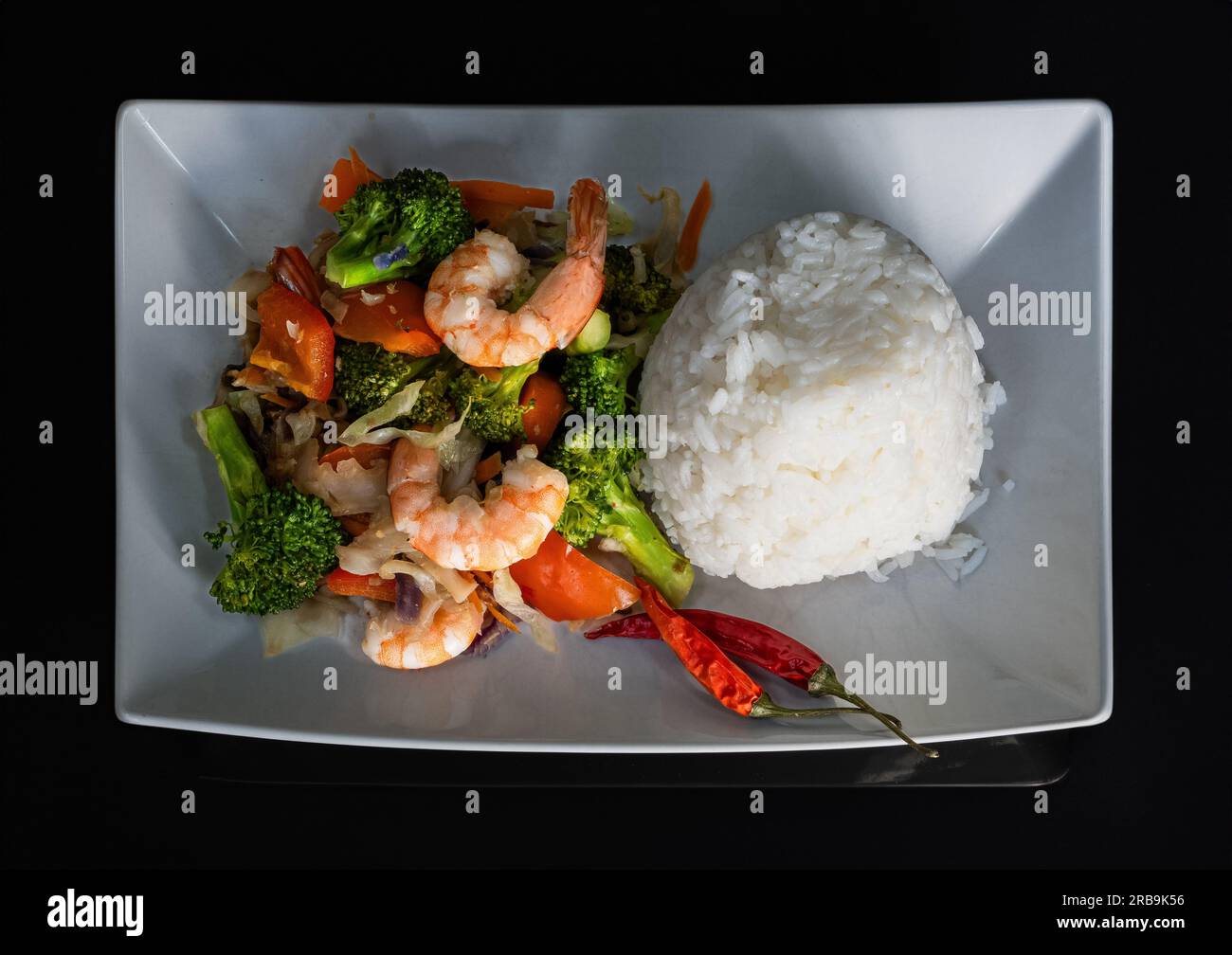 On a striking black background, a white plate showcases a delightful, colorful, and healthy stir fry seafood dish. The exquisite combination of rice, Stock Photo