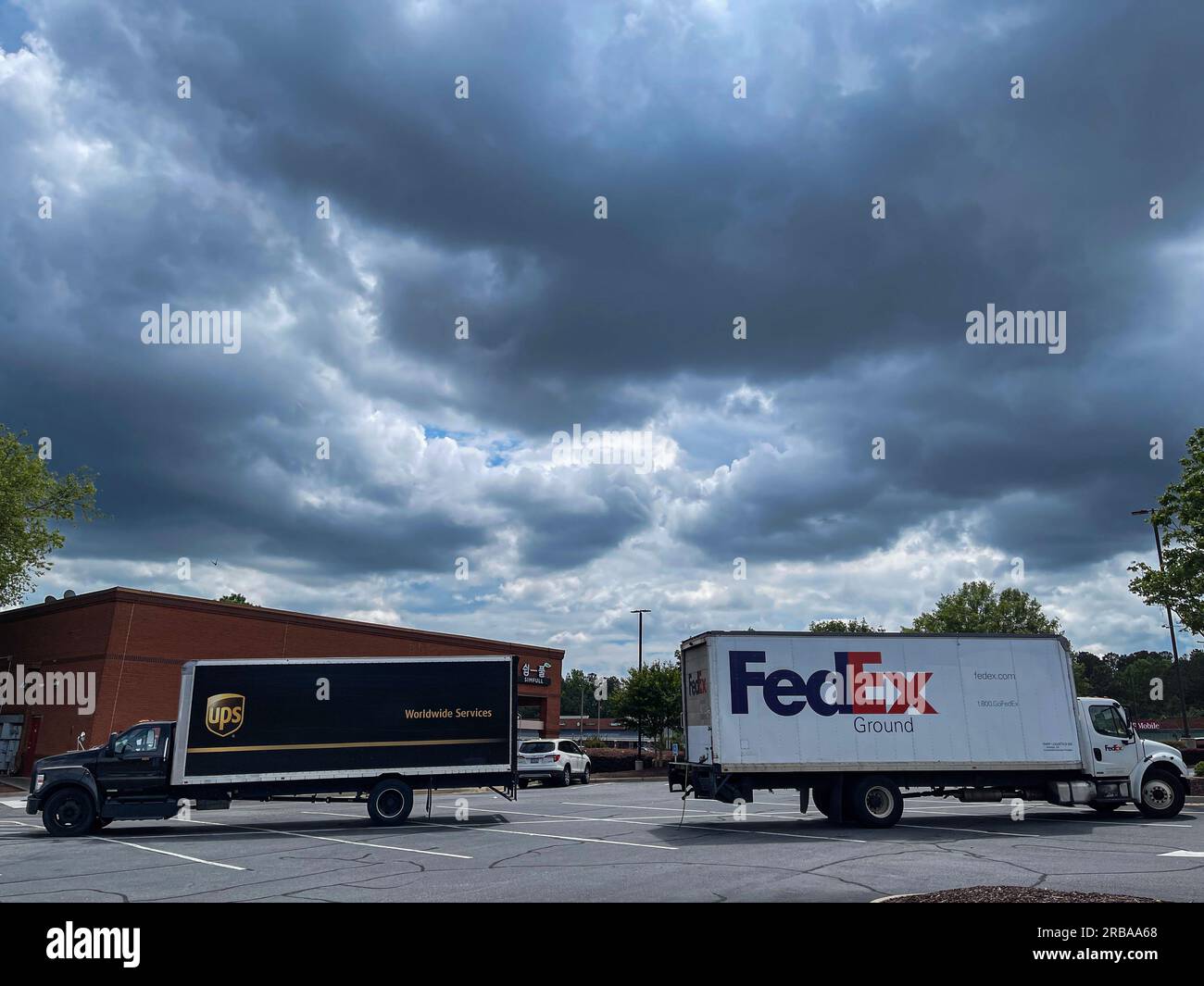 Suwanee, GA / USA - May 17, 2023:  A UPS and FedEx truck are parked opposite each other in a parking lot, with storm clouds overhead, on May 30, 2023. Stock Photo