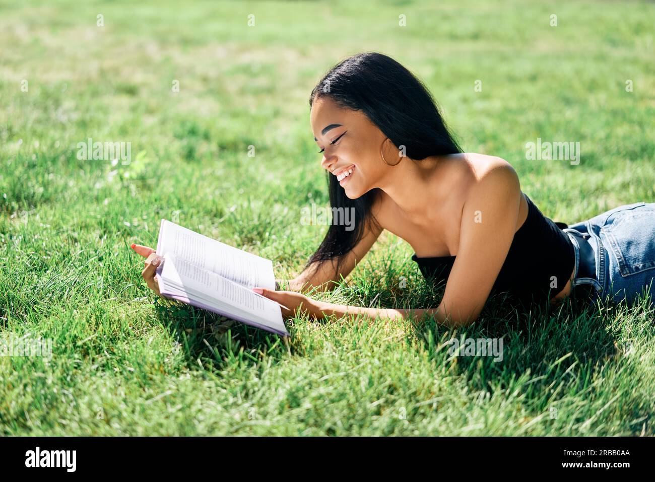 Pretty smiling afro american woman reading book lying in grass in park. Relax and hobby concept Stock Photo