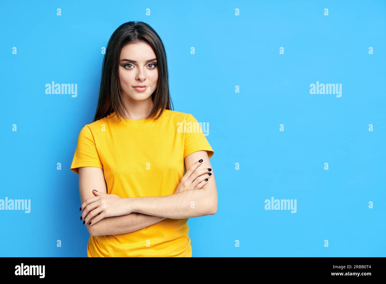 Pretty confident young beautiful woman in casual clothing with crossed arms looking at camera over blue background Stock Photo