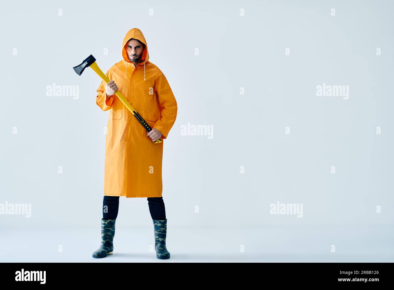 Full length portrait of young handsome man in bright raincoat holding big axe with copy space. Brutal male, lumberjack, fisherman with ax Stock Photo