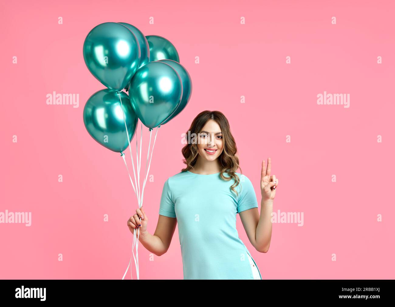 Happy pretty woman with blue balloons in hands doing victory sign on pink background. party concept Stock Photo
