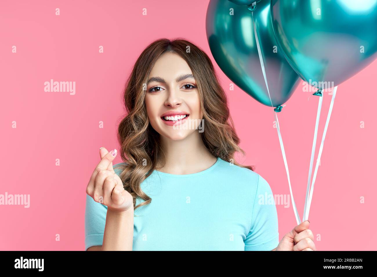 Pretty smiling woman makes mini heart korean love symbol by fingers and holding blue balloons on pink background. love concept Stock Photo