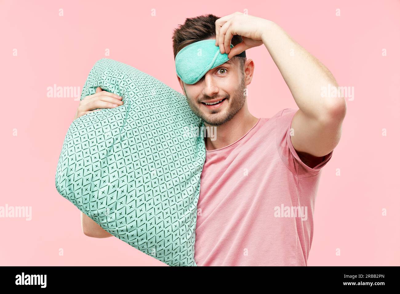 Funny cheerful man in sleeping mask holding pillow in hands looking to camera over pink background. Relax, good morning concept Stock Photo