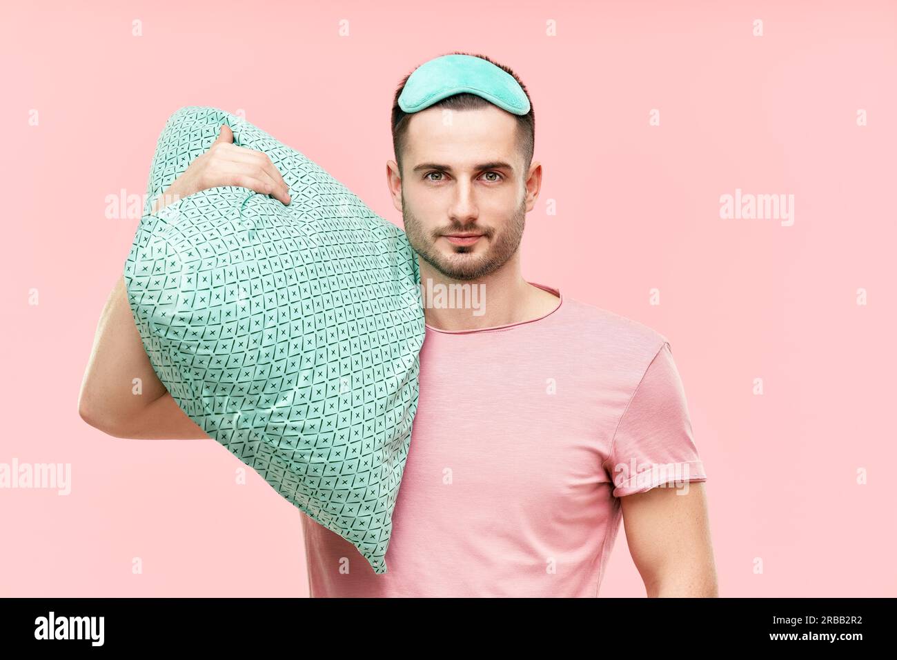 Young handsome man in sleeping mask holding pillow in hands looking to camera over pink background. Relax, tired concept Stock Photo