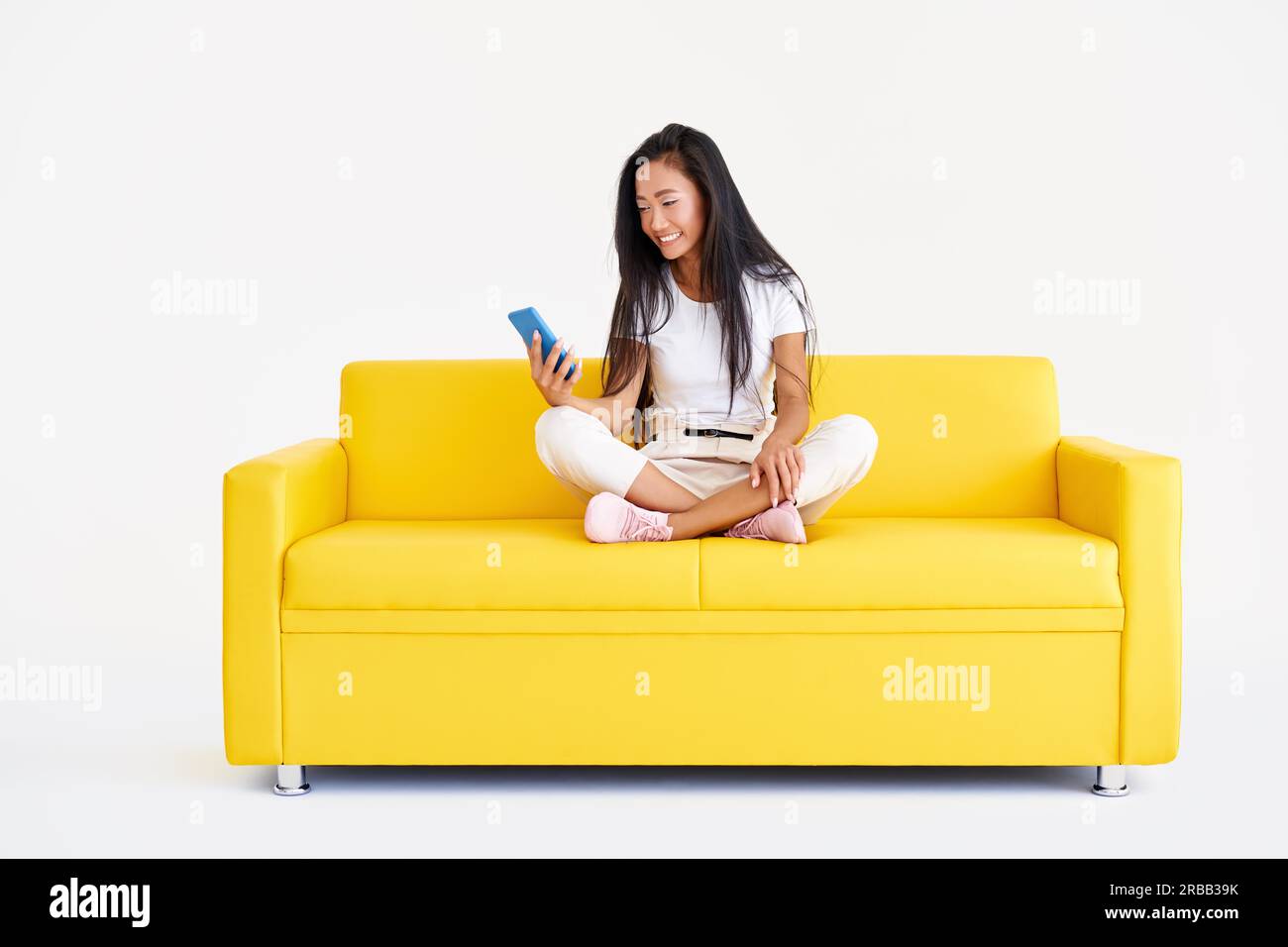 Smiling asian woman holding smartphone browses internet and scrolls news sitting on yellow couch on white background. Technology, communication Stock Photo