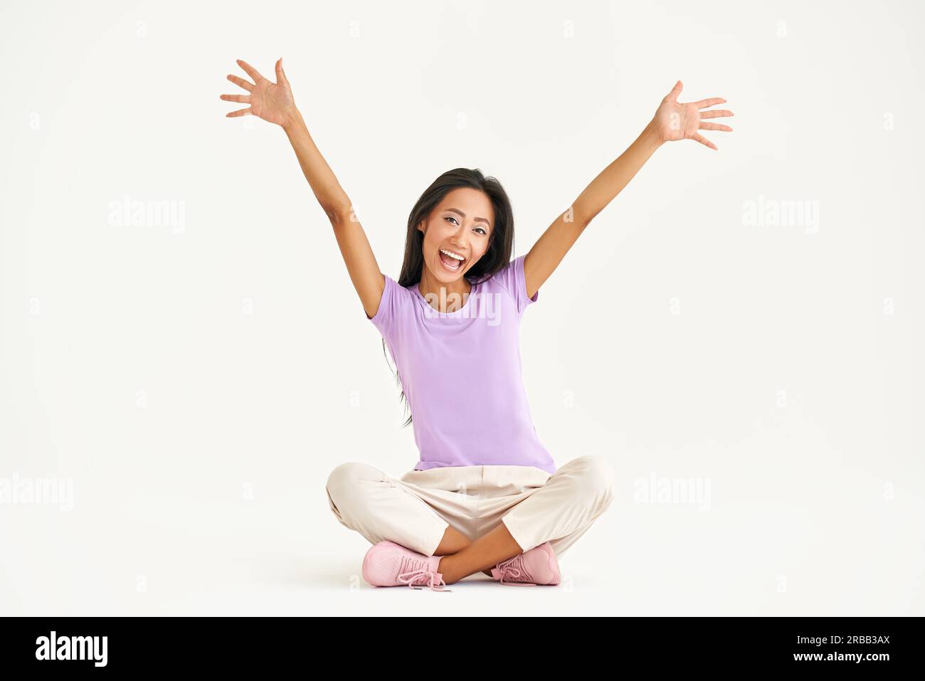 Smiling young asian woman raised arms up screaming and celebrating while sitting sitting on floor with legs crossed over white background Stock Photo