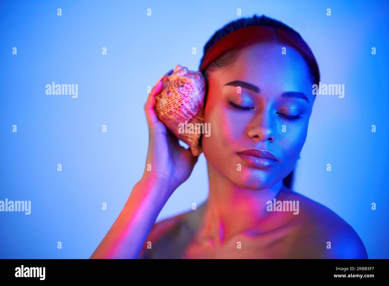 Pretty afro woman with closed eyes pressed seashell to her ear enjoy and dreams with copy space in neon light. Summer vacation, relaxation concept Stock Photo