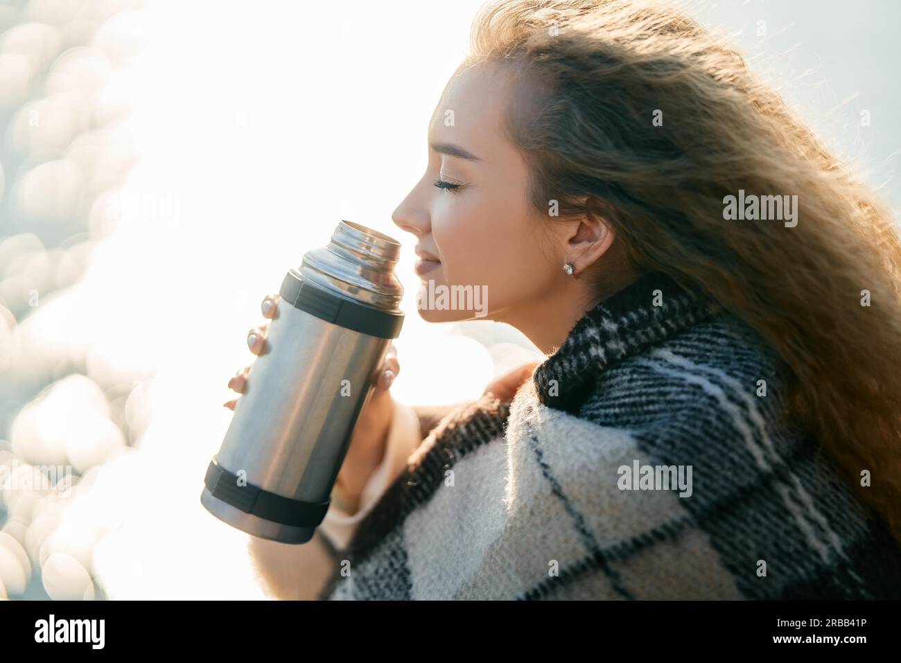 Young pretty woman holds a thermos and drinks tea wrapped in blanket over blur river background. Relax concept, happy autumn Stock Photo