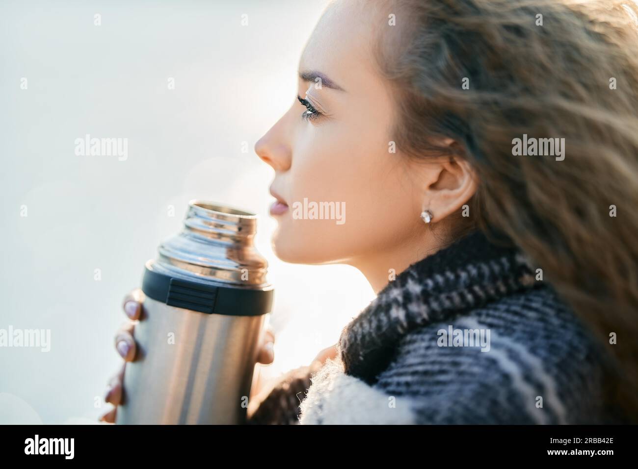 Young pretty woman holds a thermos and drinks tea wrapped in blanket over blur river background. Relax concept, happy autumn Stock Photo