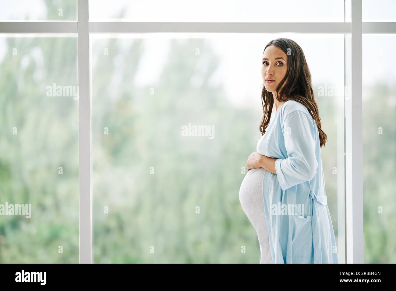 Portrait of young pregnant woman standing near window holding hands on