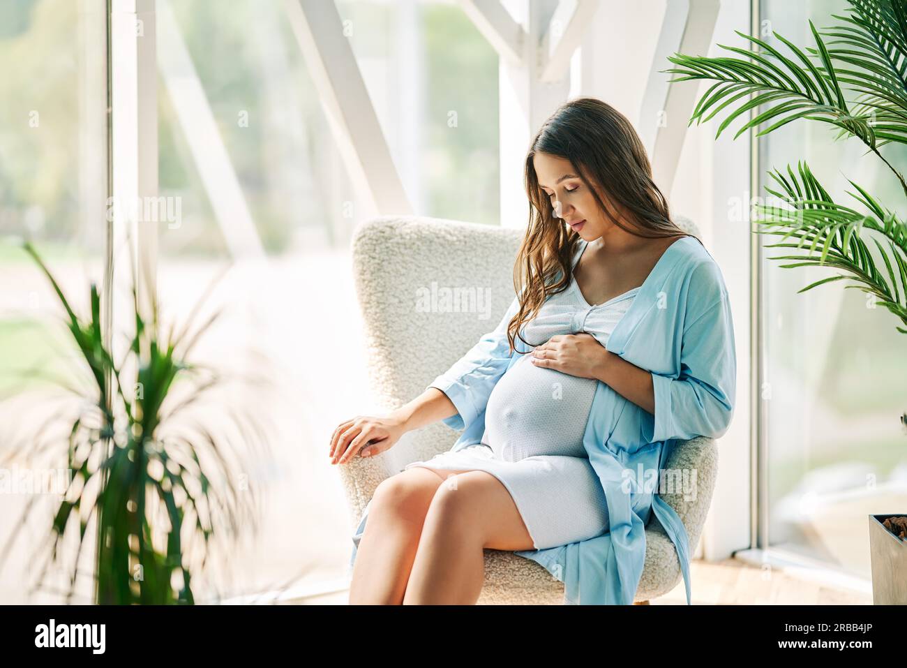 Pregnant happy woman touching her belly rest on chair at modern home. Tender mood photo of healthy pregnancy Stock Photo