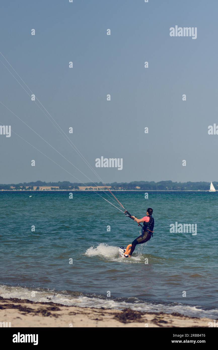 Brunette woman kitesurfing or kite boarding pulling away from the sandy beach making for deeper water on a sunny summer day in a rear view to the Stock Photo