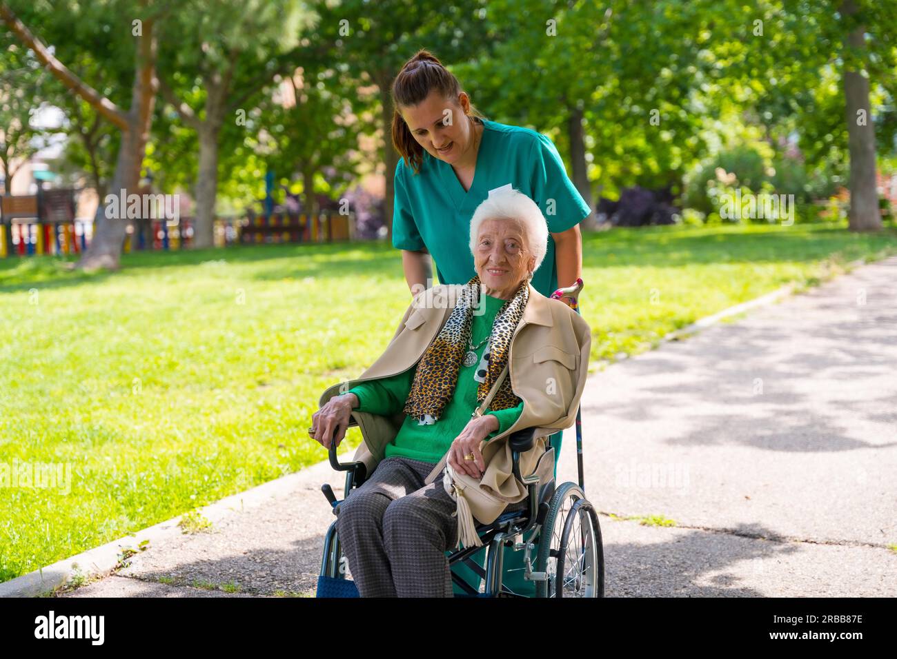 An elderly woman with the nurse on a walk through the garden of a nursing home in a wheelchair next to nature and trees Stock Photo