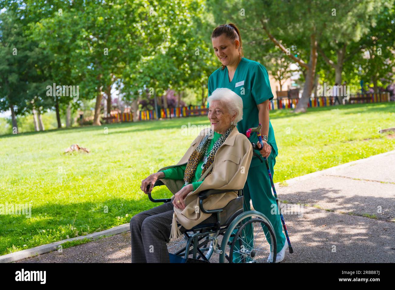 An elderly woman with the nurse on a walk through the garden of a nursing home in a wheelchair next to nature and trees Stock Photo