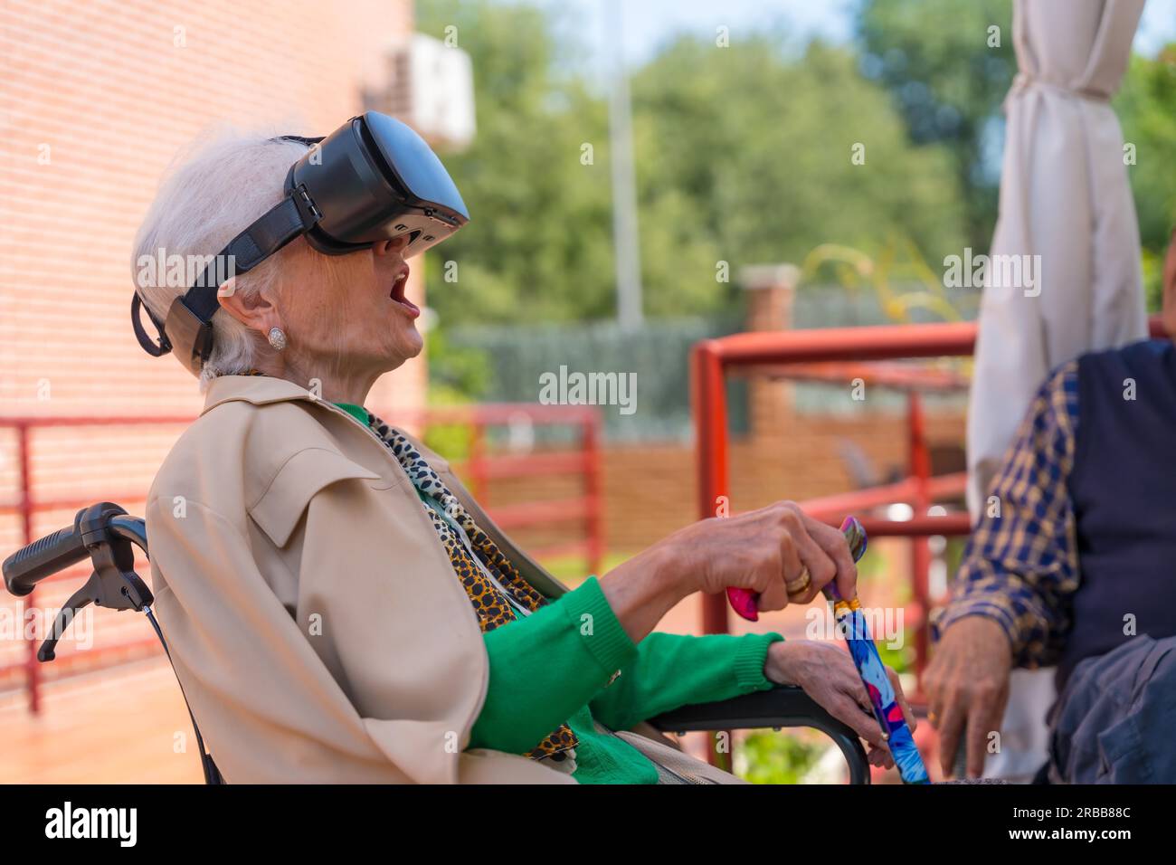 An elderly woman with the nurse looking through virtual reality glasses in the garden of a nursing home, vr glasses with an open mouth Stock Photo