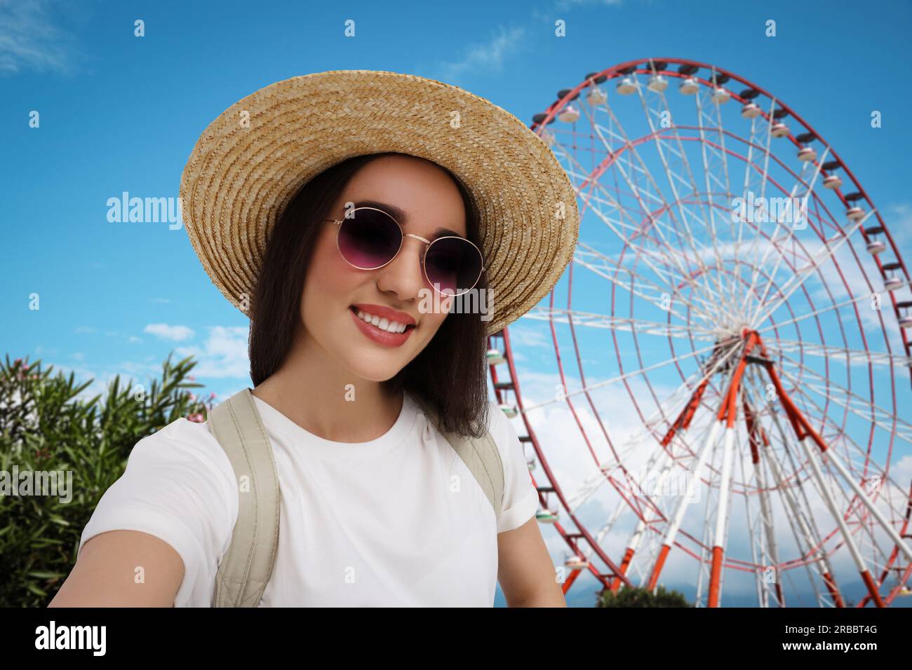 Smiling young woman in sunglasses and straw hat taking selfie near observation wheel Stock Photo