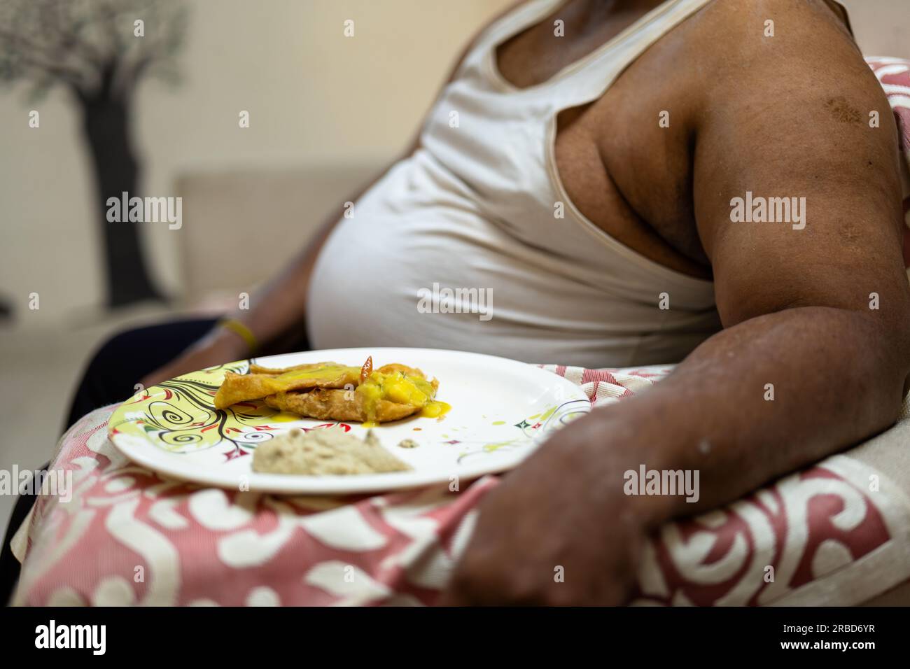 A stock photo of a fat man sitting at a sofa, focusing on a plate of food. The man's belly is out of focus, Stock Photo