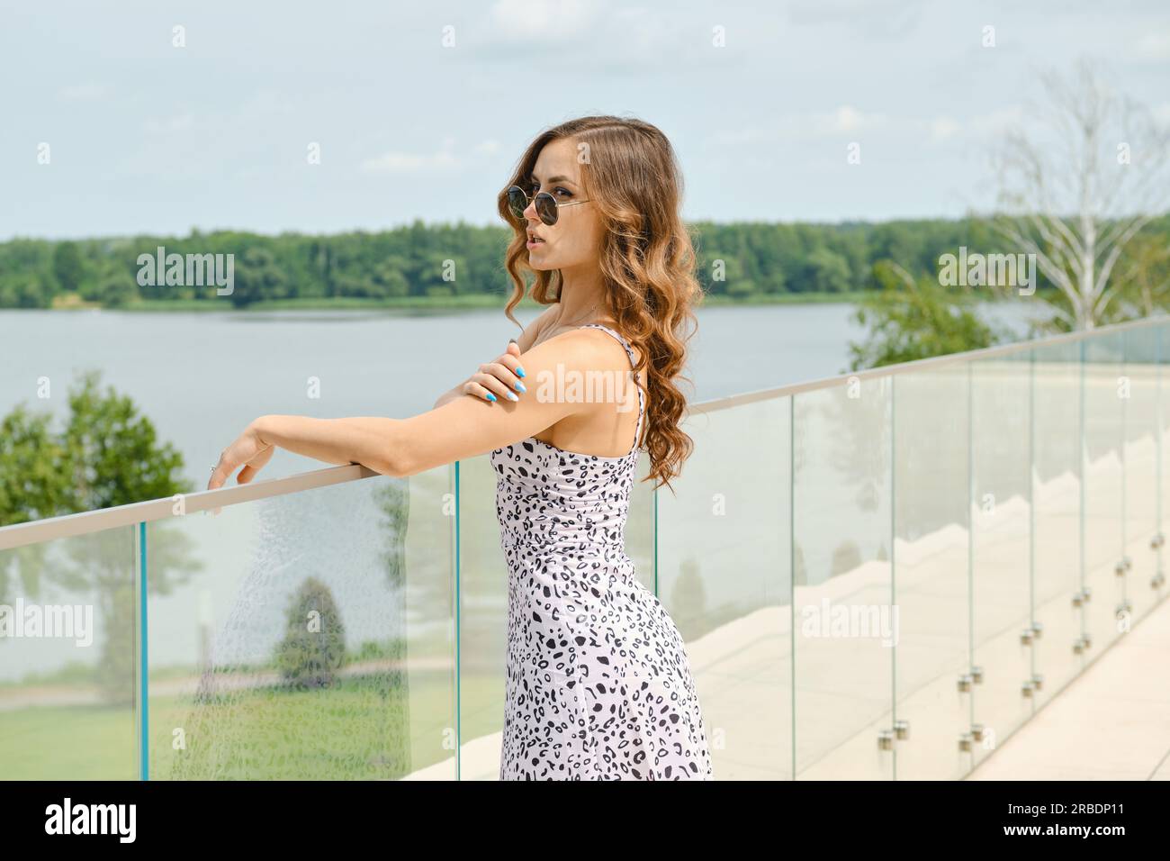 Young woman in sundress and sunglasses looking at water standing on balcony of hotel Stock Photo