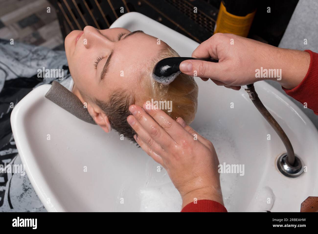 The barber's hands pour rain on the client's head and wash their hair with water in the sink, before cutting their hair at the hairdresser. Stock Photo