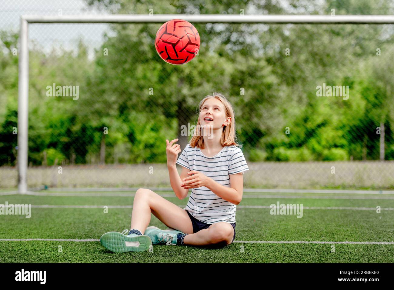 Pretty child girl sitting on grass and throwing football ball up. Cute