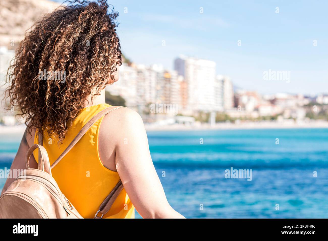 Seen from back, unrecognizable caucasian curly hair woman dressed in yellow contemplating a city nest to sea. Stock Photo